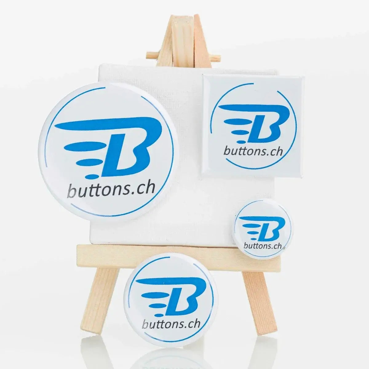 Runde und quadratische Metallbuttons mit blauem buttons.ch-Logo auf einer kleinen Staffelei.