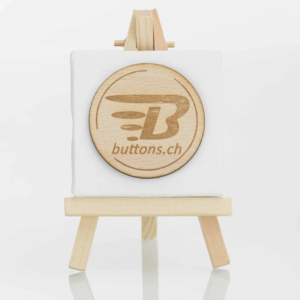 Runder Holz-Anstecker, 56 mm, mit eingraviertem buttons.ch-Logo auf einer kleinen Staffelei.
