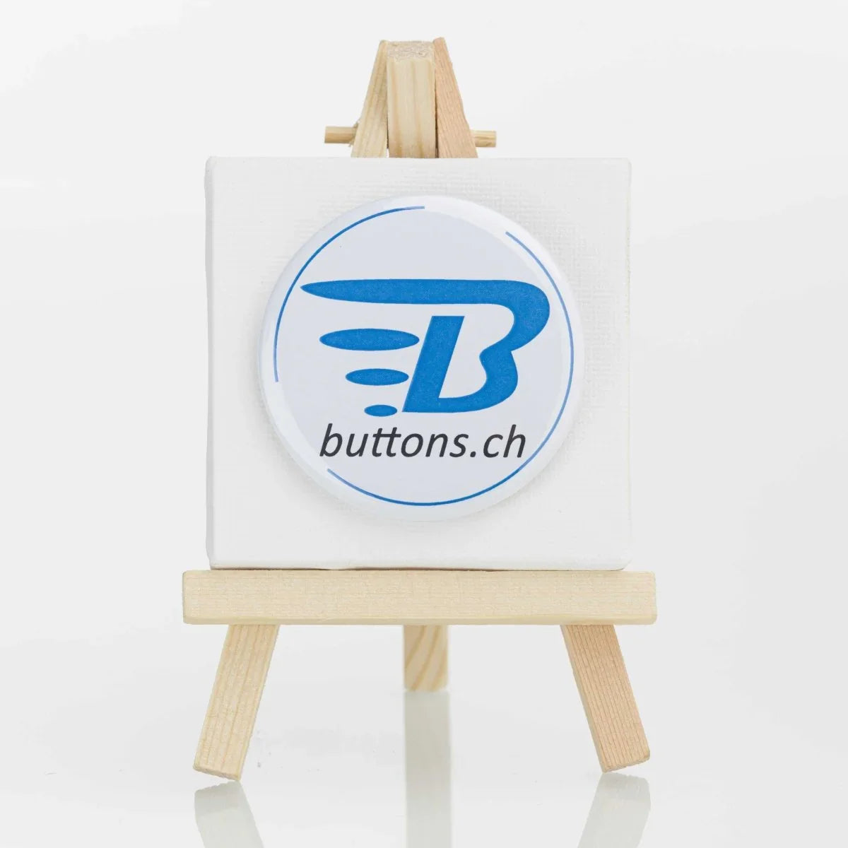 Runder Metallbutton, 56 mm, mit blauem buttons.ch-Logo auf einer kleinen Staffelei.